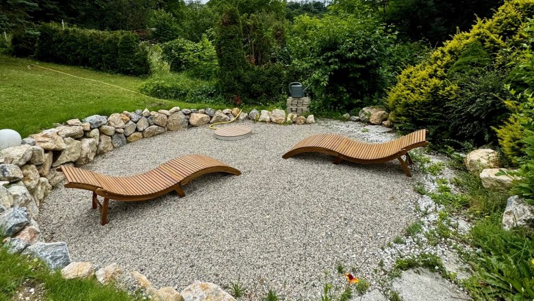 Two wooden loungers on a gravelled area, surrounded by stones and bushes, under a cloudy sky.