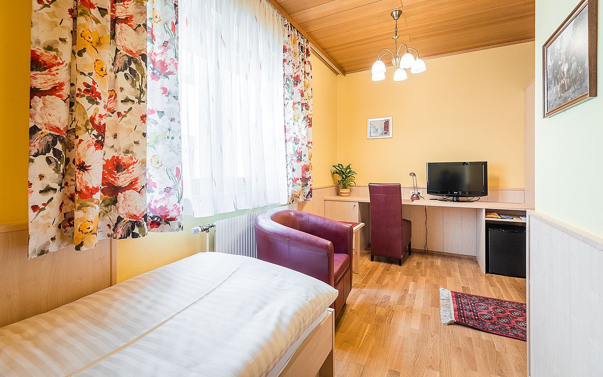 A cozy single room with a wooden floor, a bed, a desk with chair, a TV and colorful curtains.