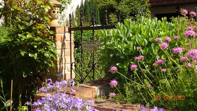 A garden with a small gate and a sign saying 'I'm in the garden'. Surrounded by flowering plants and a brick post.