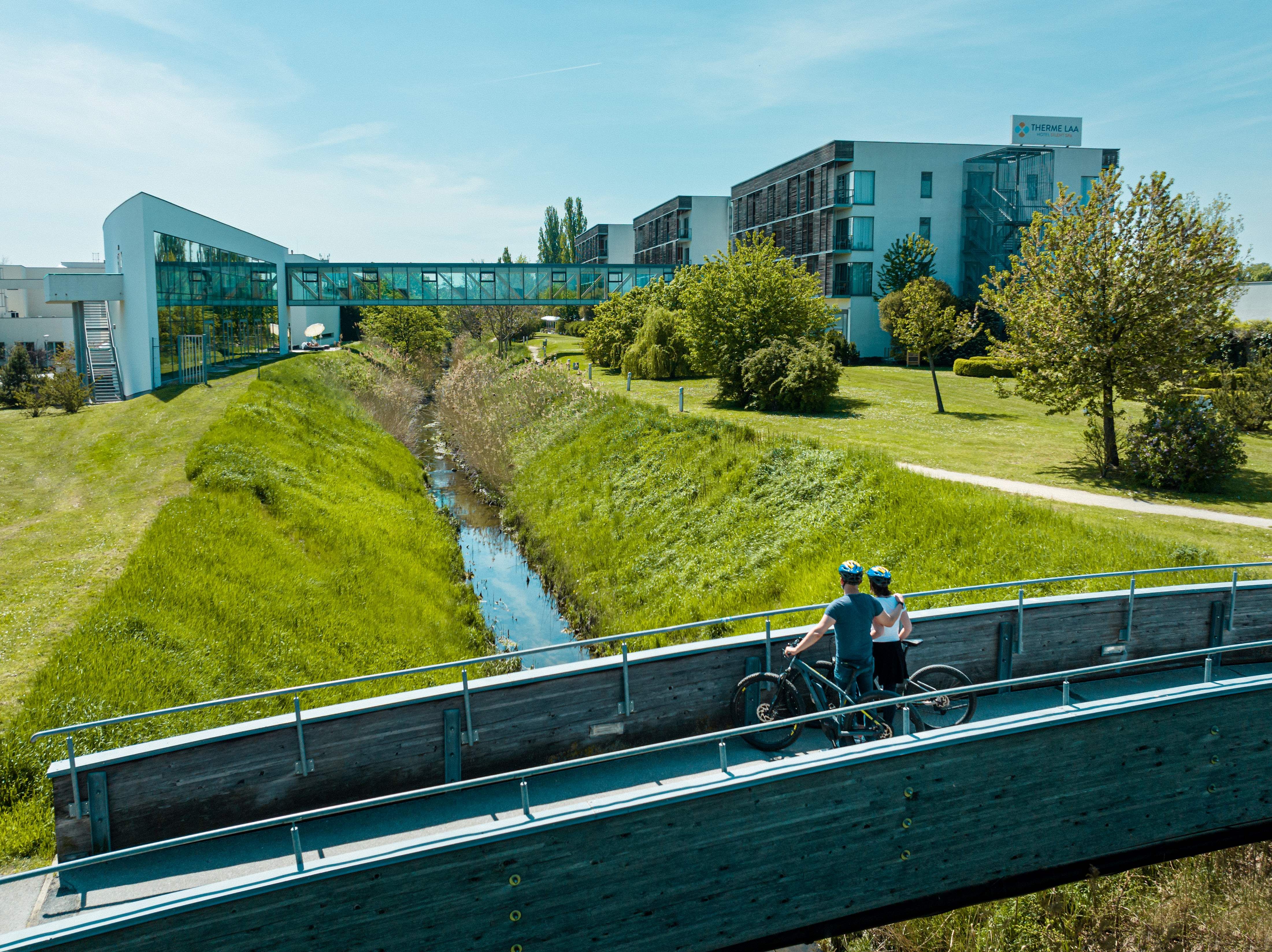 Two cyclists on a bridge in front of the Therme Laa & Hotel.