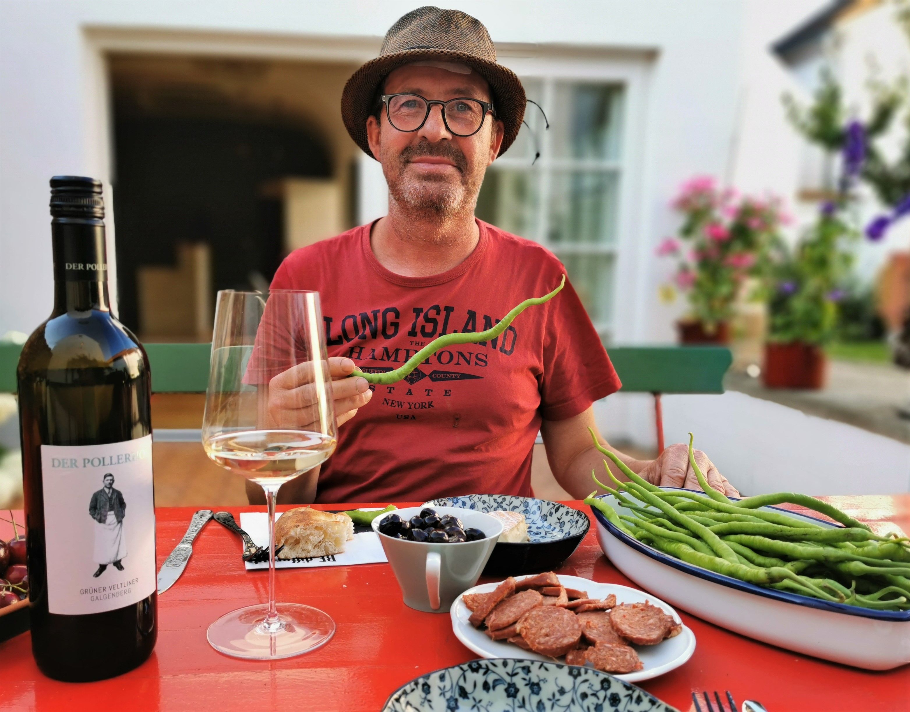 A man in a hat sits at a table with a bottle of wine, a glass, beans and sausage.