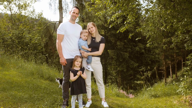 A family is standing in a meadow in front of a forest. A man, a woman, a small child in the woman's arms and another child are standing together.