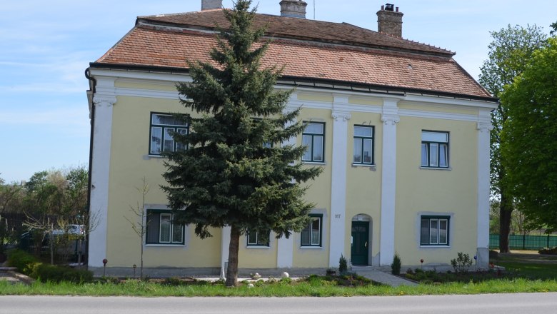 Yellow two-storey building with red tiled roof and green door, surrounded by trees and a small garden.
