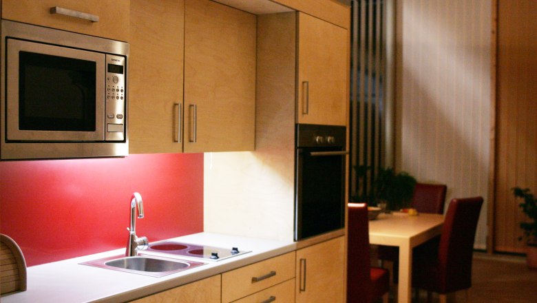 Modern kitchen with wooden cupboards, red splashback, microwave and dining table in the background.