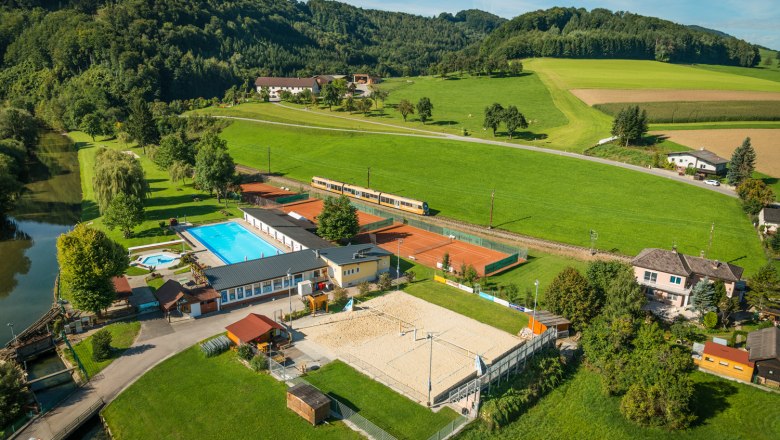 Aerial view of an outdoor swimming pool with pool, tennis courts and volleyball court, surrounded by green fields and hills.