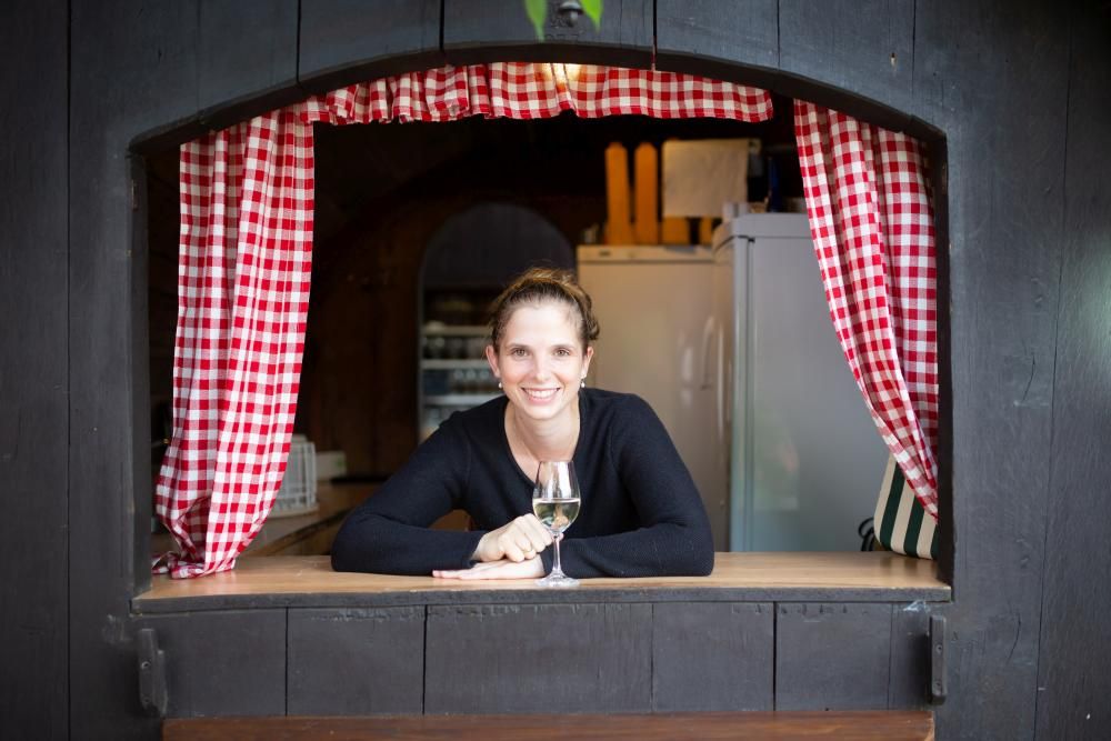 Woman leaning out of a window with red and white checkered curtains, holding a glass of wine.