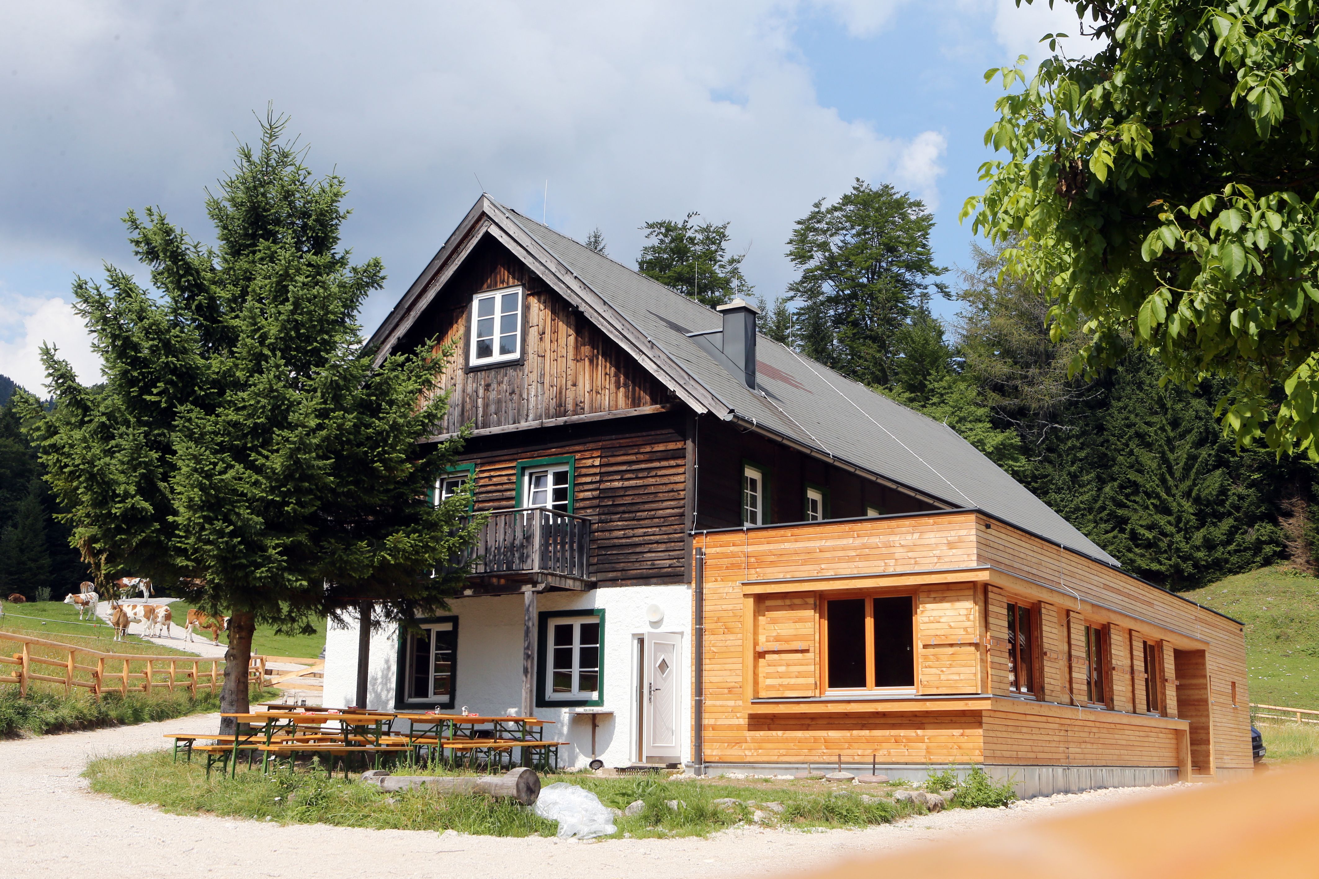 A traditional wooden house with extension in a rural setting, surrounded by trees and meadows.