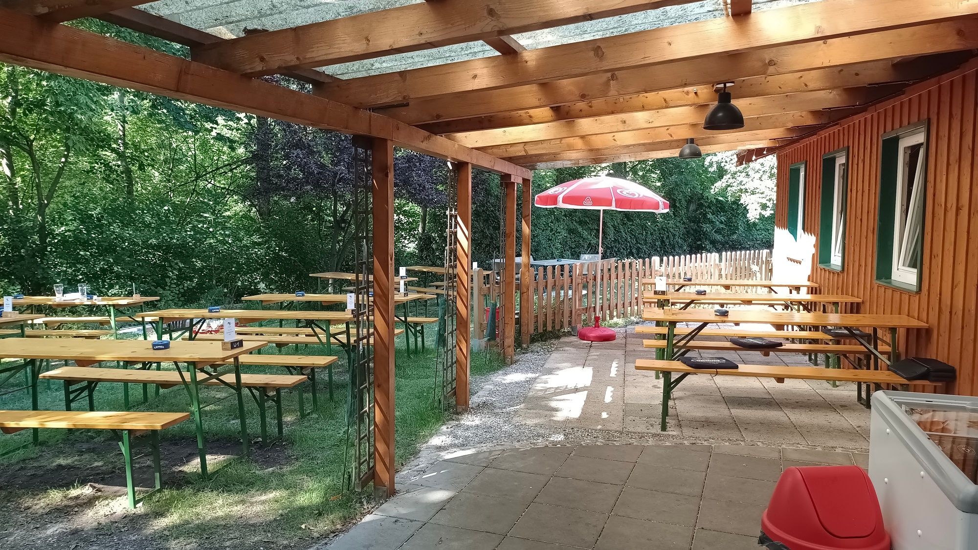 Covered terrace with wooden benches and tables, surrounded by trees and a wooden fence.