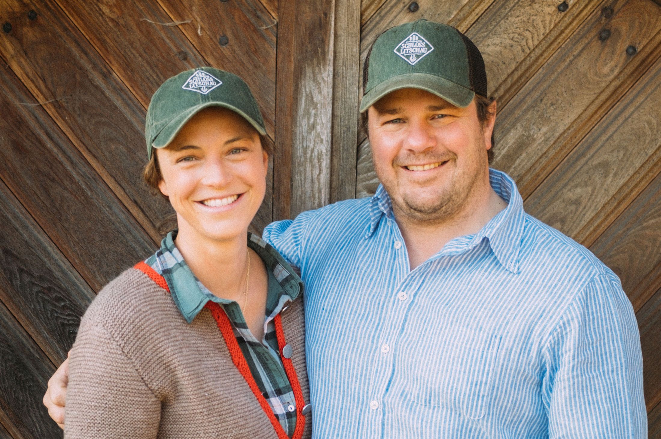 Two people are smiling at the camera, wearing green caps with a logo and standing in front of a wooden wall.