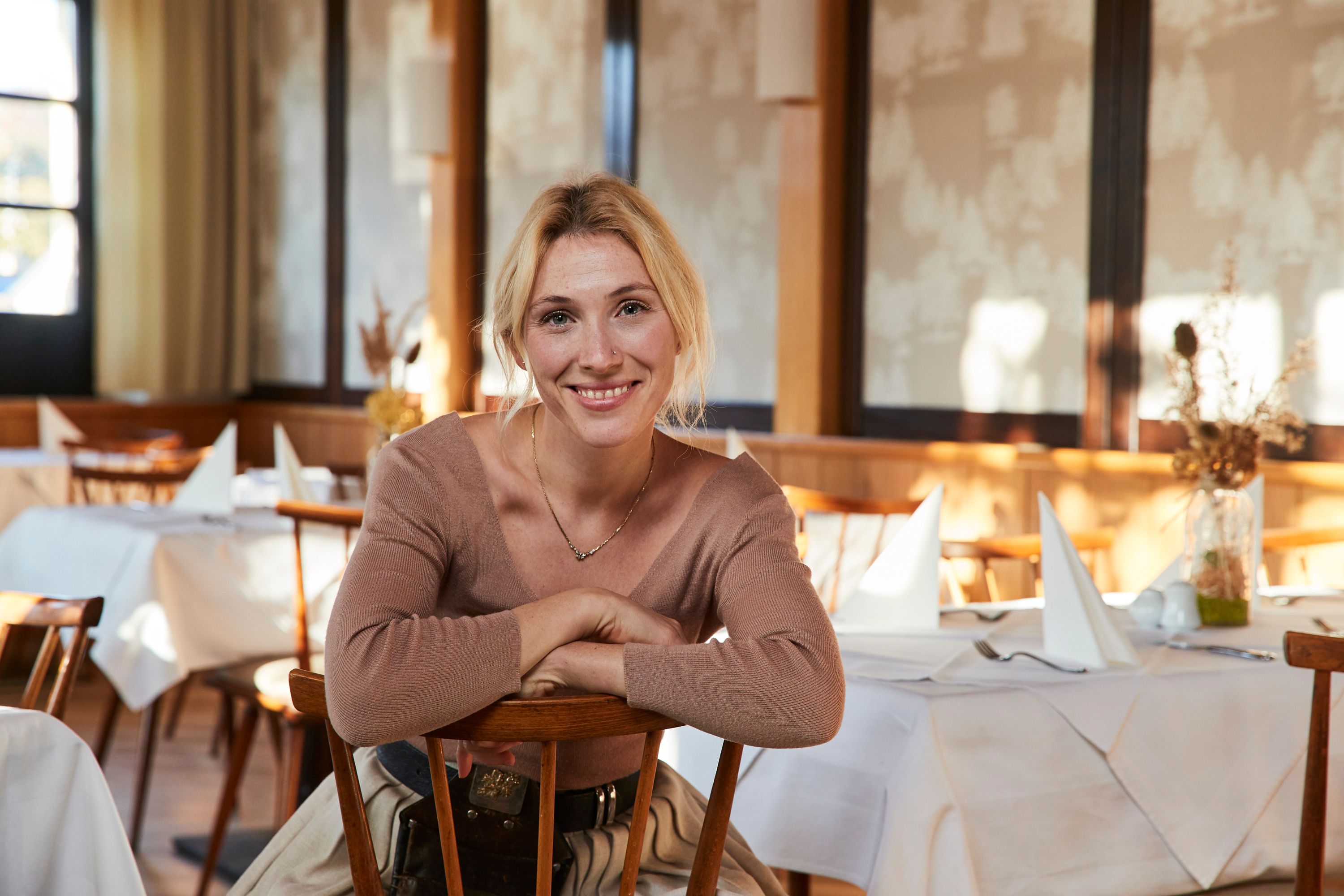 Woman sitting smiling in a restaurant with set tables.