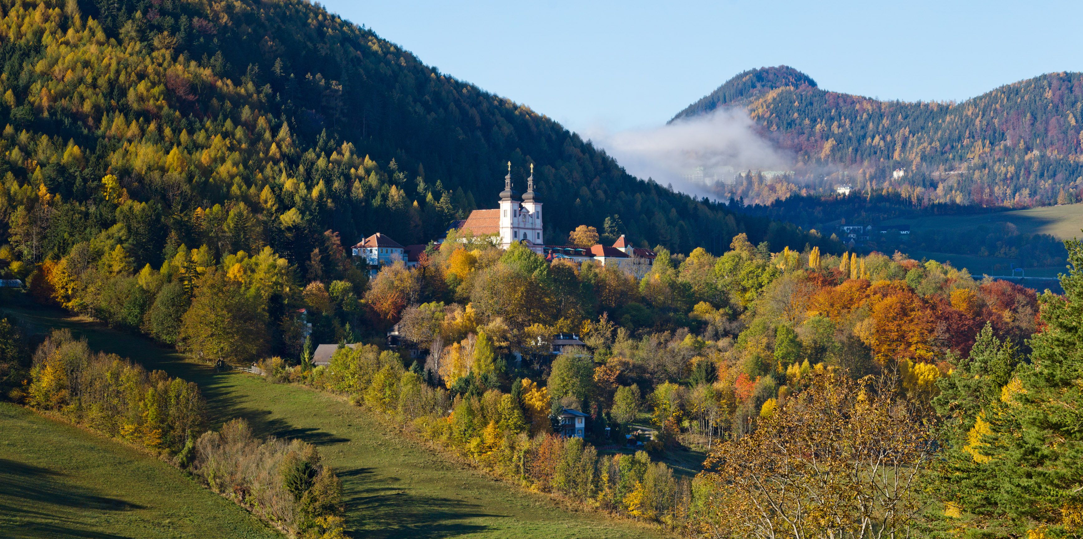 Autumn landscape with church in Maria Schutz, surrounded by colorful trees and hills.