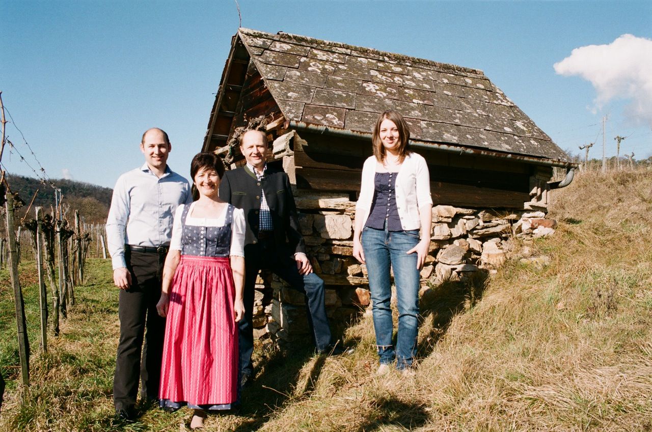 Four people are standing in front of an old hut on a hill with vines in the background.