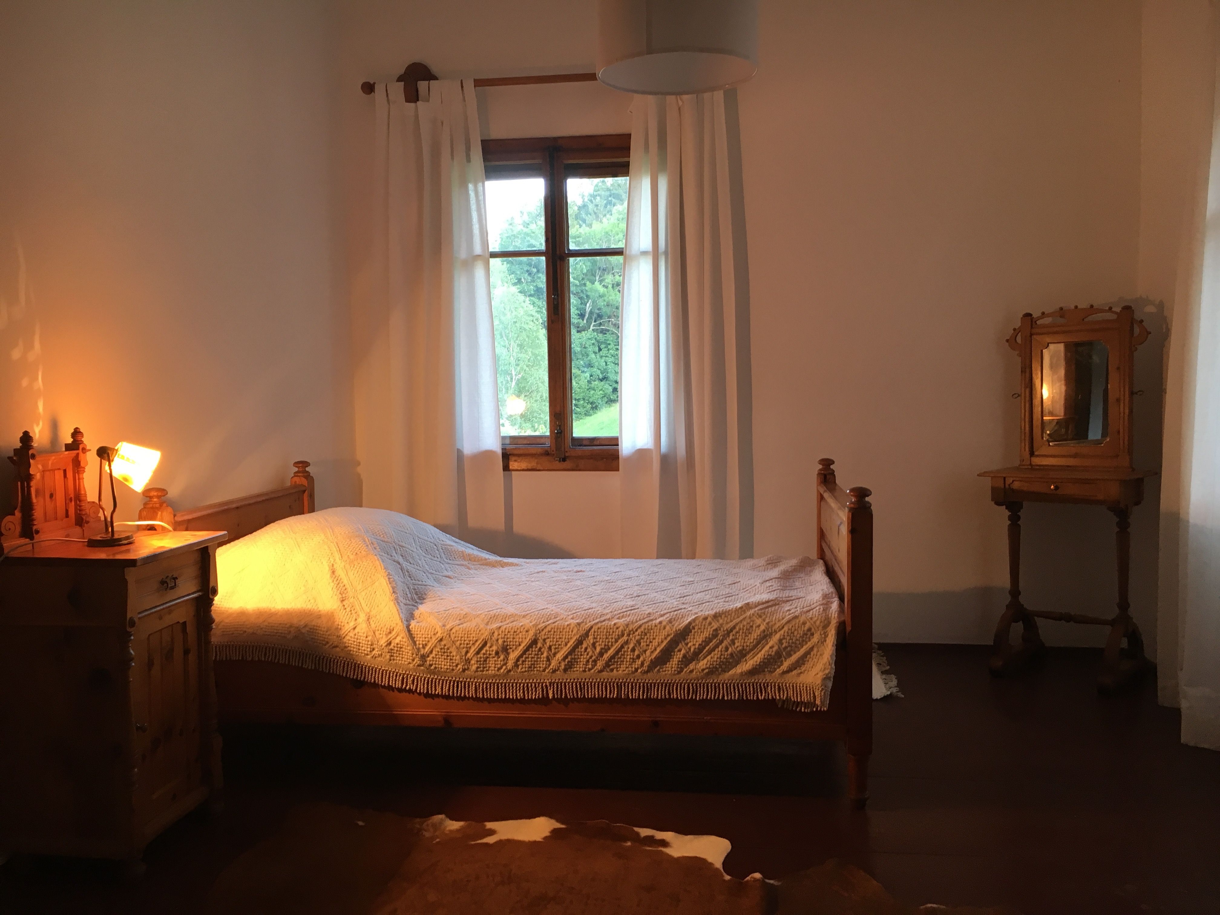 A rustic single room with wooden furniture, a bed, a bedside table with lamp and a window with white curtains.