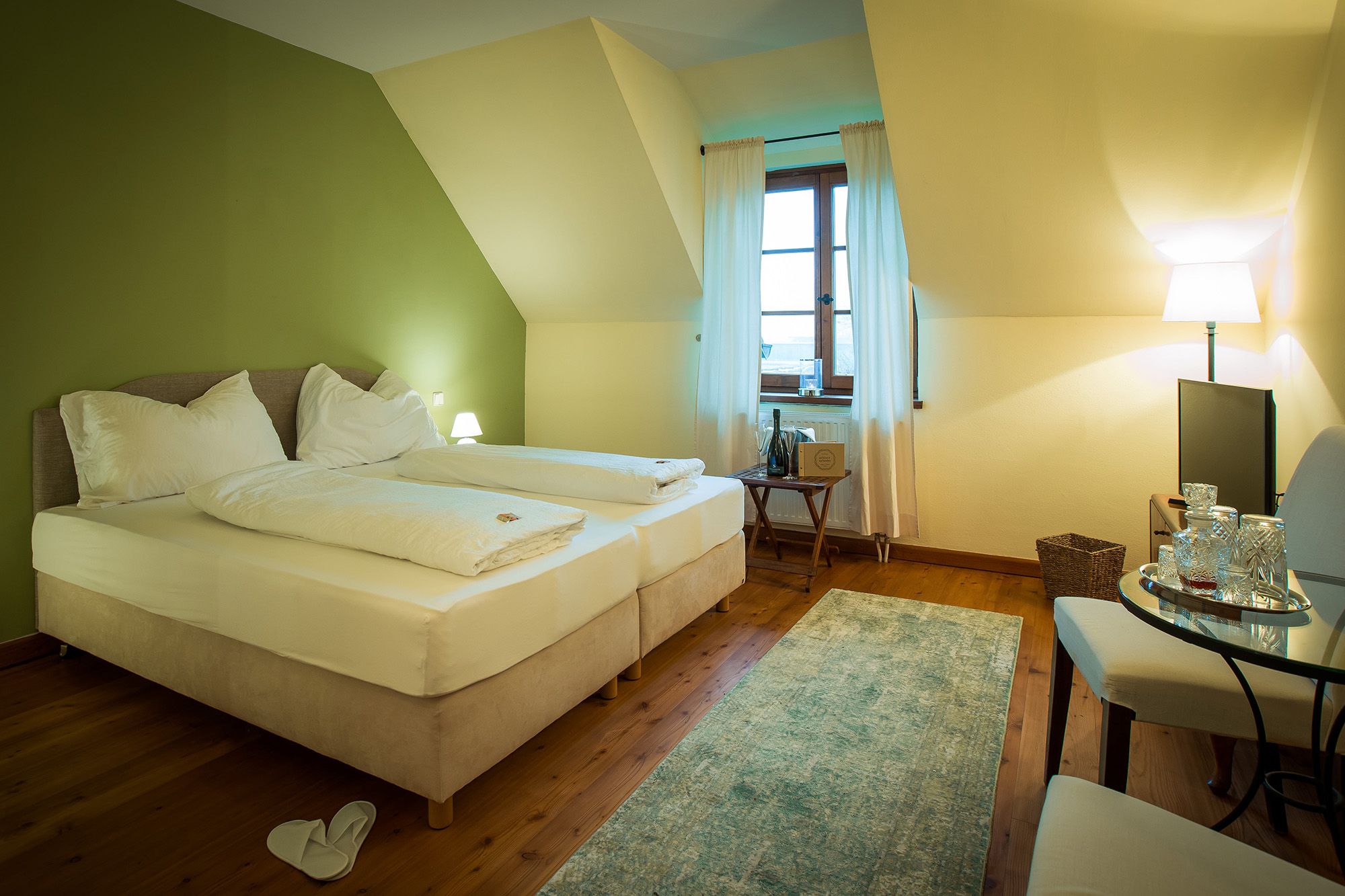 A cozy double room with a green-painted wall, two beds, a window with curtains, a small table and a TV.
