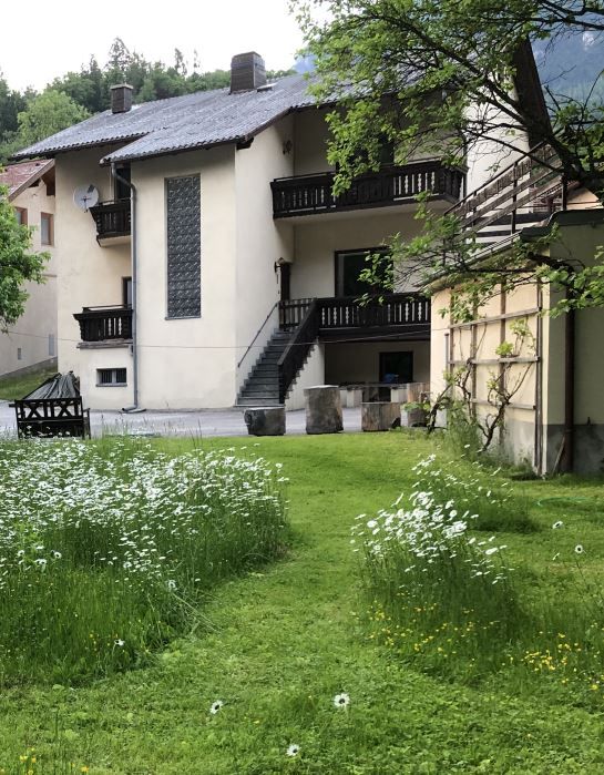 A two-storey house with a balcony and stairs, surrounded by a green garden with flowers.