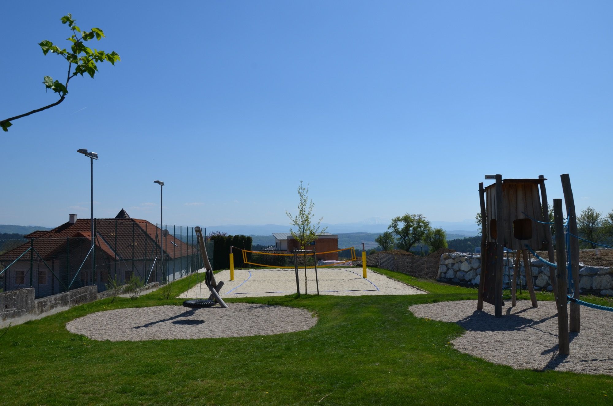 A beach volleyball court with a net, surrounded by green grass and a playground in the foreground, under a clear blue sky.