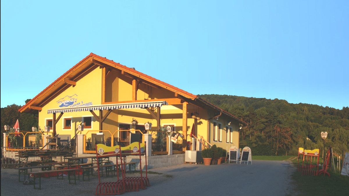 A yellow building with a terrace and beer garden furniture, surrounded by green hills under a clear sky.