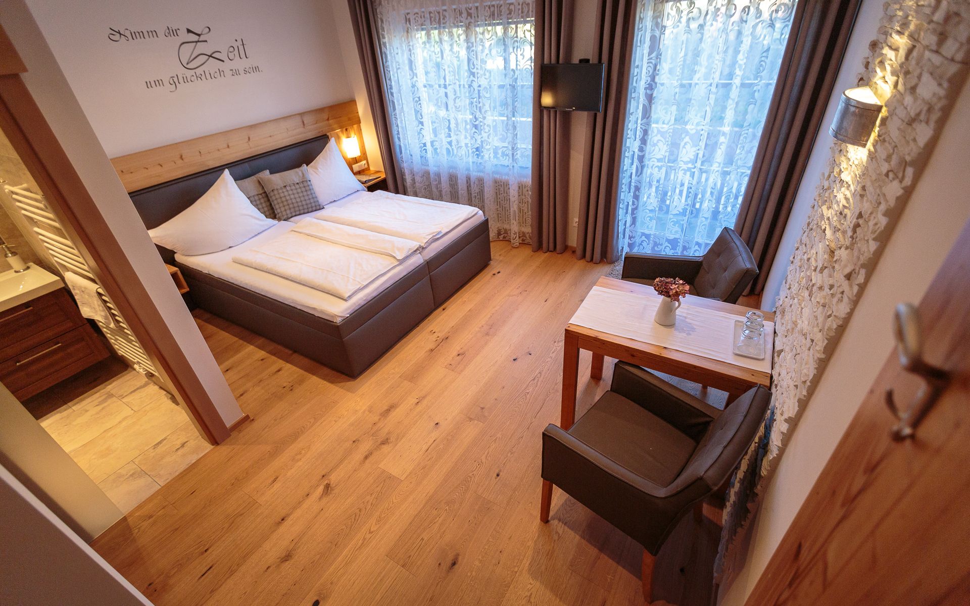 Cozy double room with wooden floor, double bed, small table and chairs, decorative curtains and a TV on the wall.
