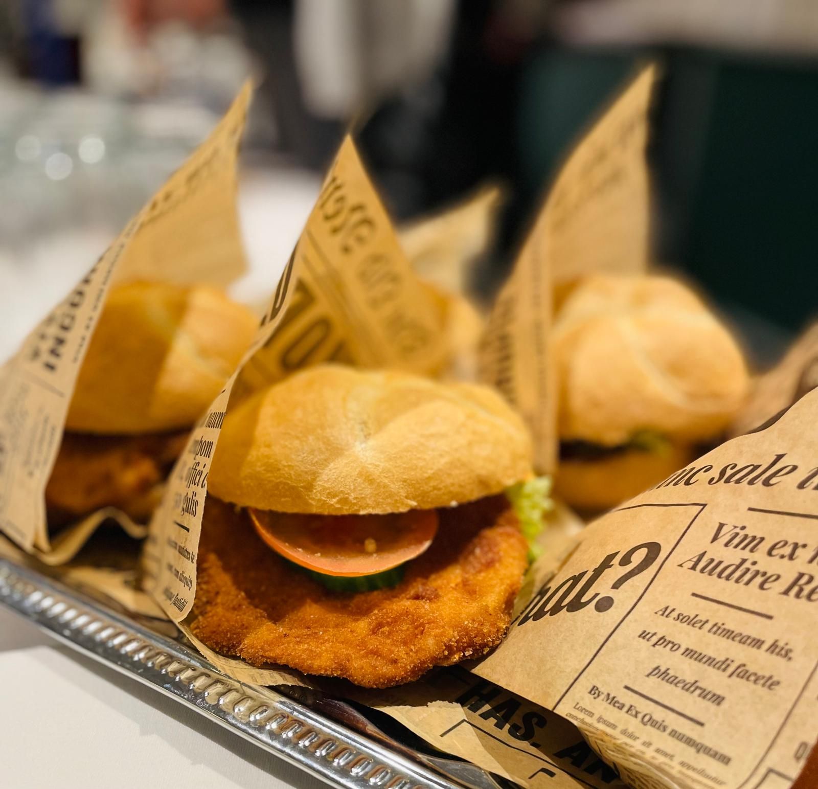 Several burgers wrapped in newspaper on a tray; in the foreground, a burger with a breaded patty, tomato, and cucumber.