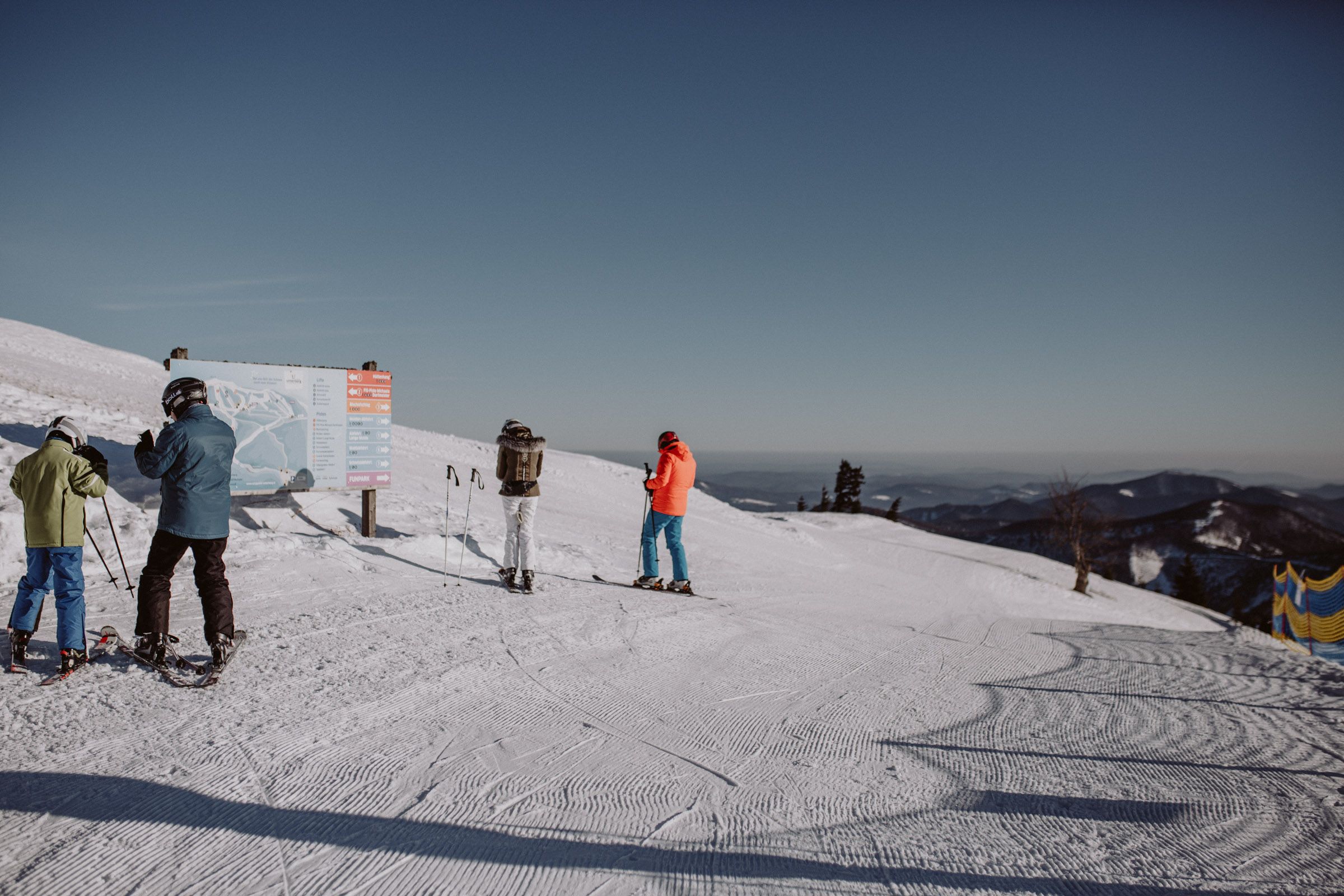 Skiers stand on a slope in the Unterberg ski area in front of a signpost, surrounded by snow-covered mountains and a clear sky.