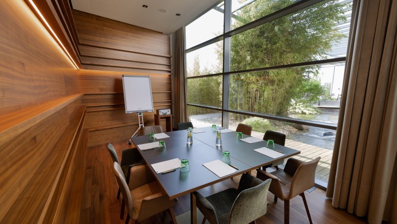 Modern seminar room with wooden walls, large table, chairs and flipchart. Large windows with a view of the garden.