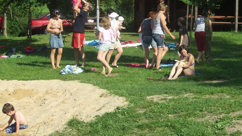 People relax by a natural bathing pond with a parasol and trees.