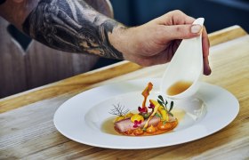 A plate with artfully arranged food, over which a hand pours sauce from a small jug.