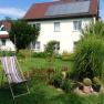 A house with solar panels on the roof, surrounded by a garden with plants and a sun lounger.