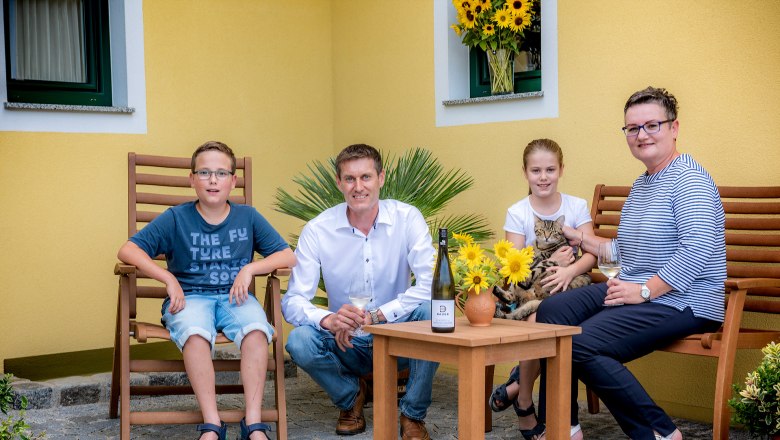 A family is sitting outside on wooden furniture in front of a yellow house. A man, a woman, a boy and a girl with a cat can be seen.