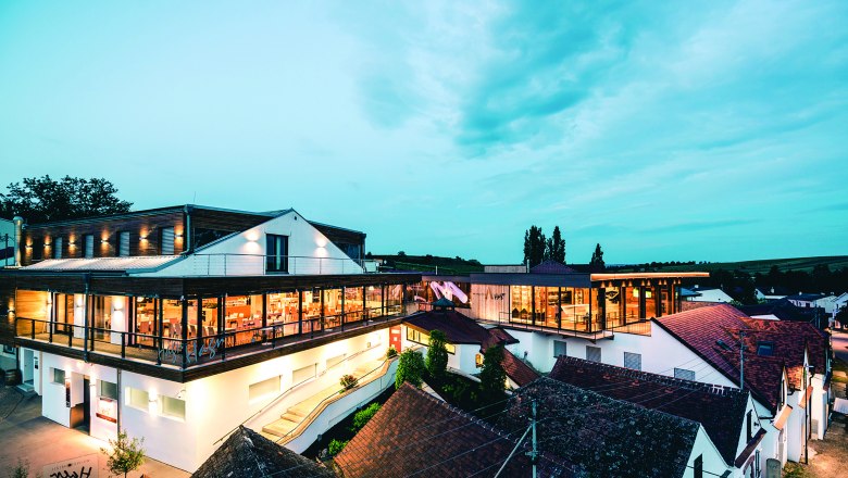 Wine domicile Hagn, © Weingut Hagn / Michael Reidinger Modern winery architecture at dusk with illuminated interiors and surrounding buildings.