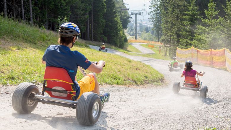 People ride down a gravel path on mountain carts, surrounded by trees and a ski lift.