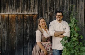 A couple is standing in front of a wooden wall, she is wearing a dirndl, he a chef's jacket.