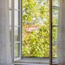 Open window with white curtains, view of green trees.