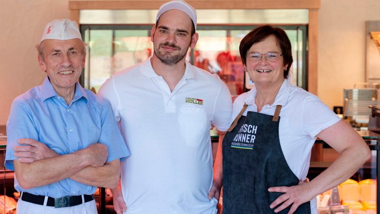 Three smiling people in work clothes are standing in a butcher's shop.