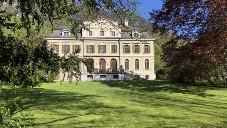 A large, elegant manor house with a symmetrical façade and several windows, surrounded by green lawns and trees.