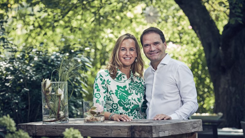 A man and a woman stand smiling at a wooden table outdoors, surrounded by green nature.