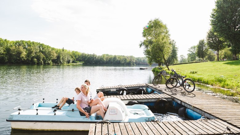 Three people on a pedal boat by the lake, bicycles on the jetty, green trees in the background.