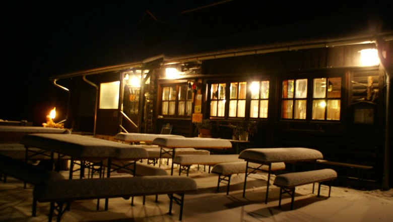 Snow-covered benches in front of an illuminated hut at night.