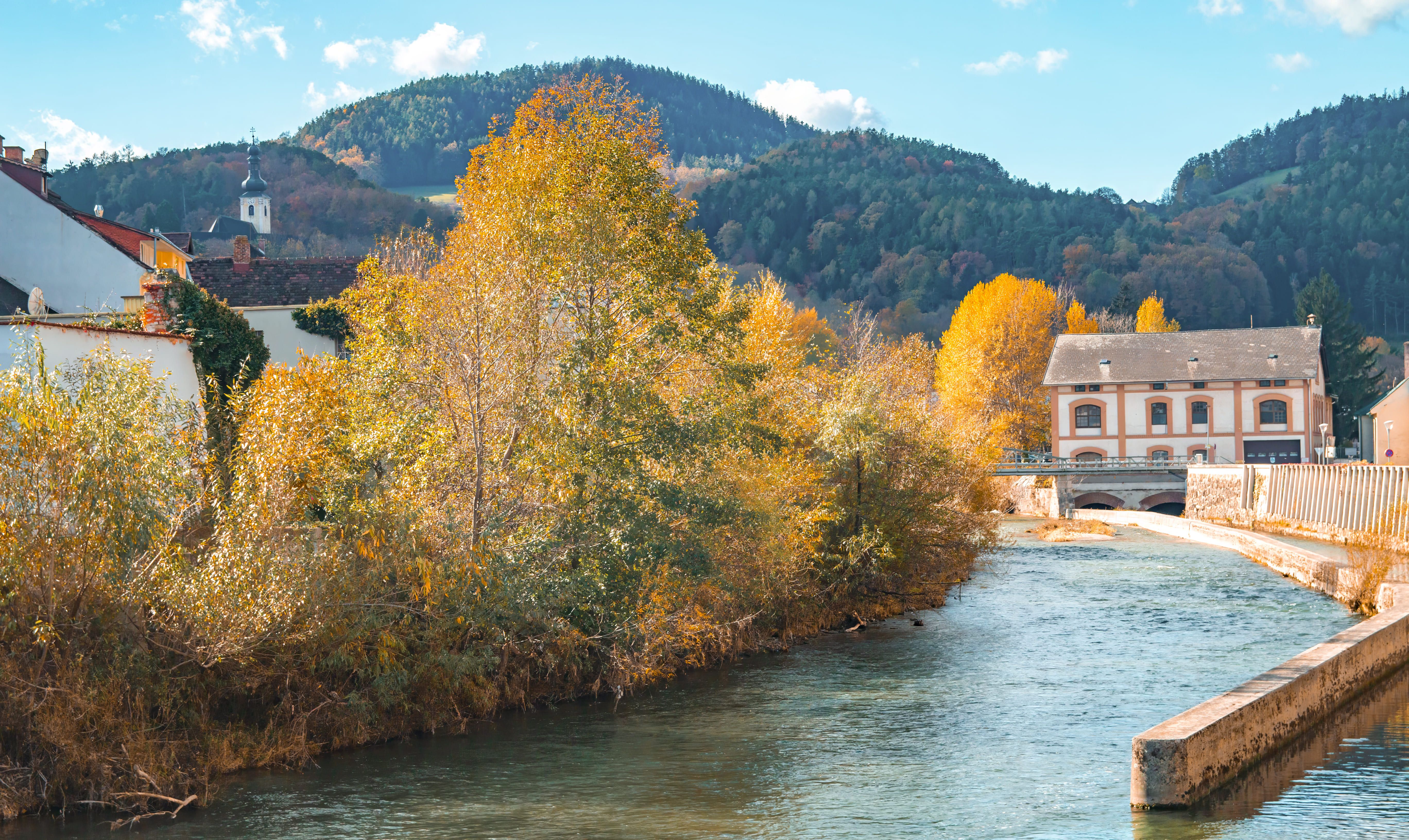 River with autumn trees and buildings in the background, surrounded by hills.