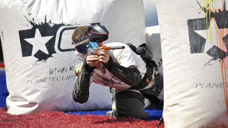 A paintball player in protective clothing aims a paintball gun behind an inflatable cover.