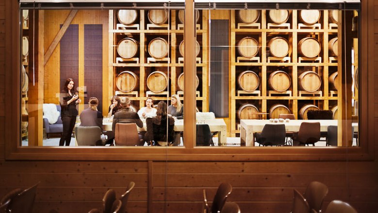 View through a window into a room with whisky barrels and a group of people sitting at a table.
