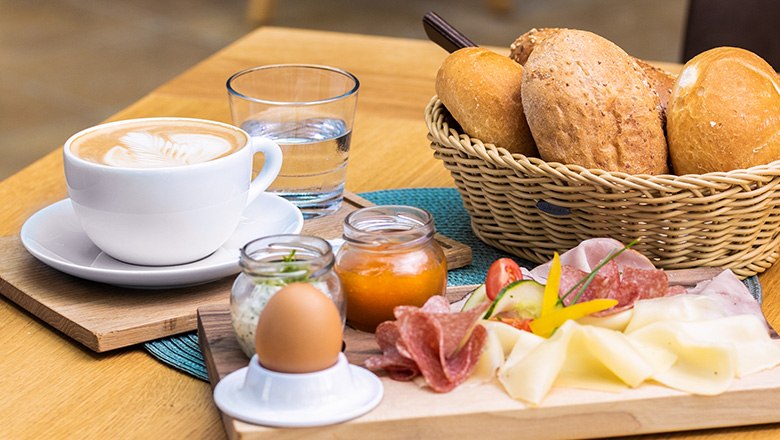 Breakfast on a wooden board, next to it a basket of pastries and a coffee with water.