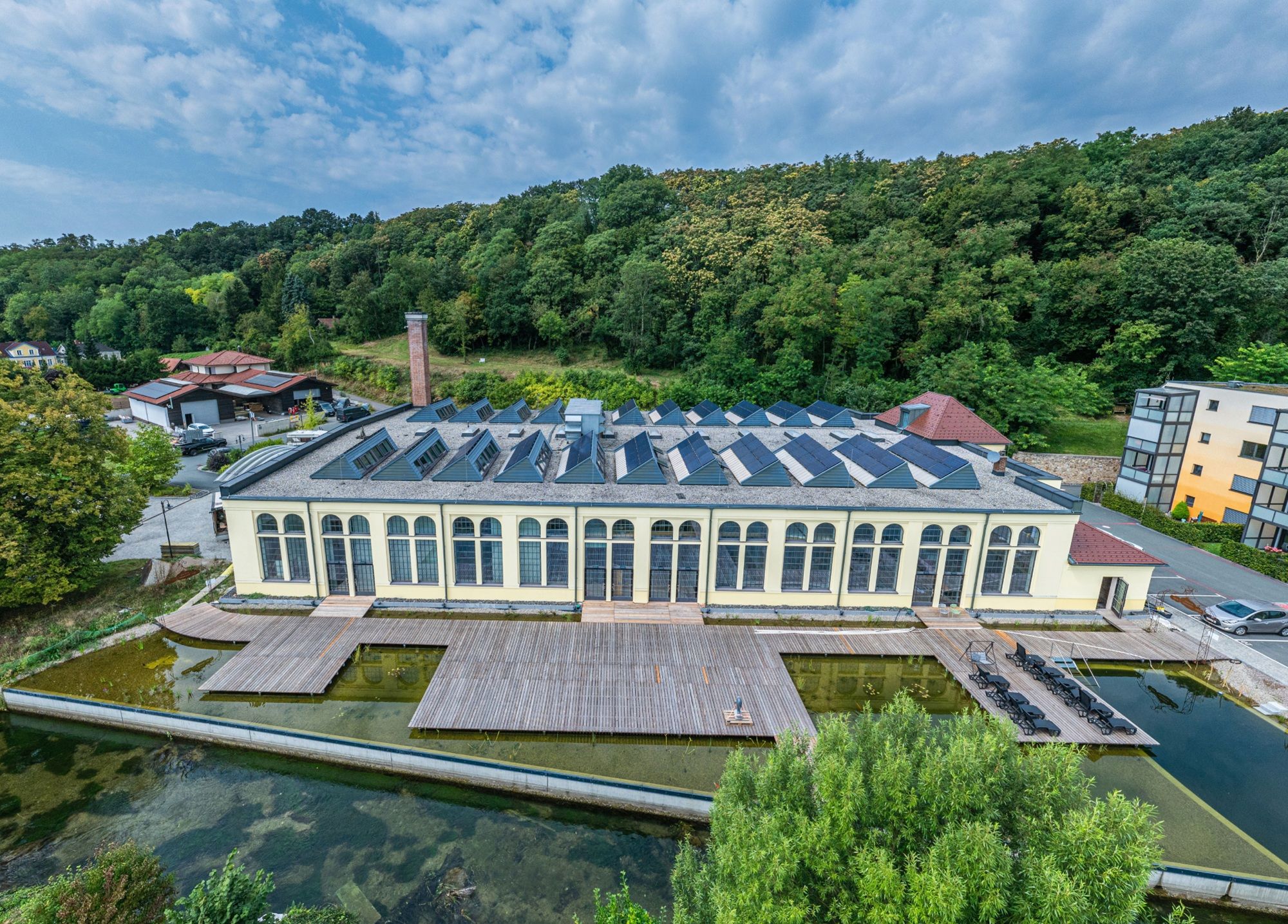 Aerial view of an old factory with solar panels on the roof, surrounded by trees and a pond.