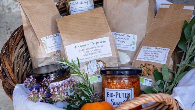 A gift basket with various products such as einkorn goods, organic turkey sugo and elderberry juice, decorated with herbs and flowers.