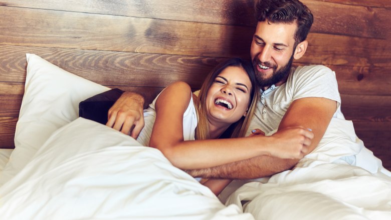A laughing couple lie together in bed in front of a wooden wall.