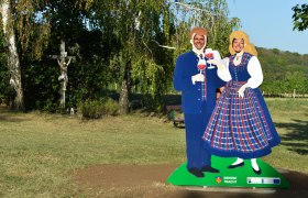 Two people pose behind a cardboard figure in traditional costume with glasses of wine. A crucifix and trees in the background.