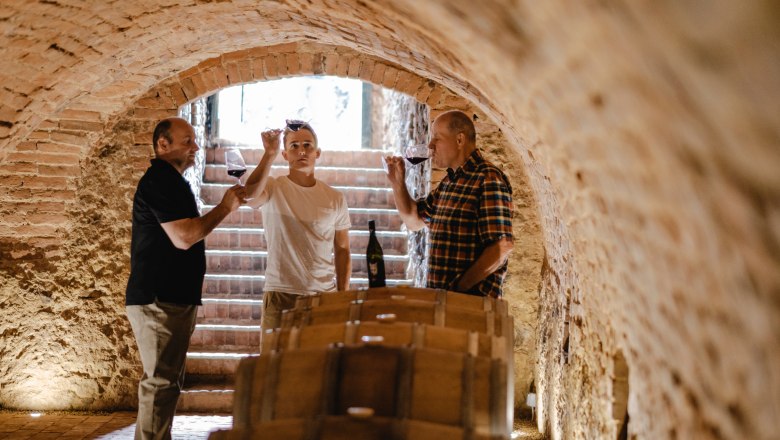Three men taste wine in a wine cellar with a brick vault.