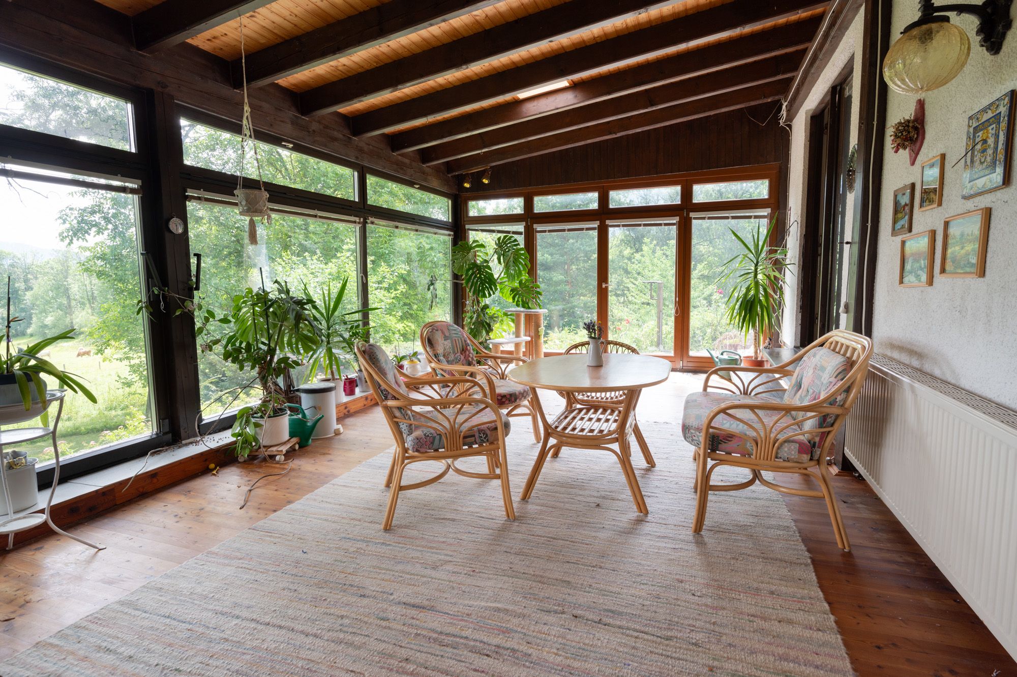 A conservatory with wooden floor, rattan furniture and lots of plants. Large windows offer a view of green nature.