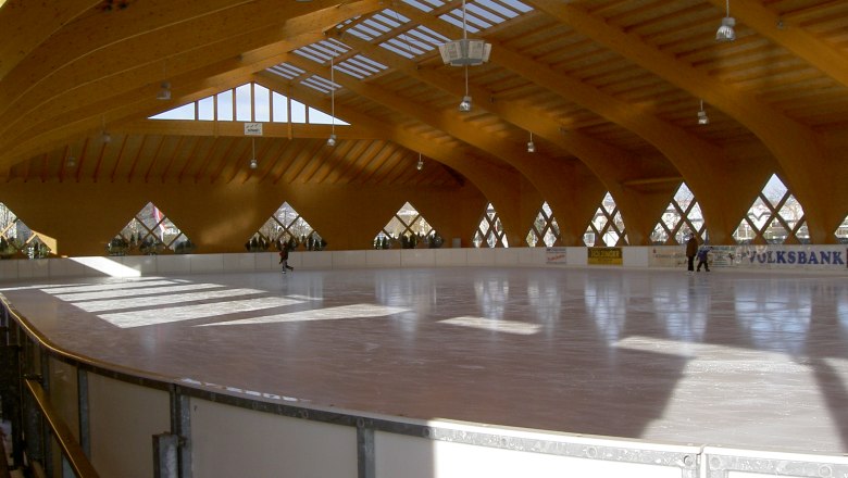 Interior view of a covered artificial ice rink with a wooden roof and few people on the ice.