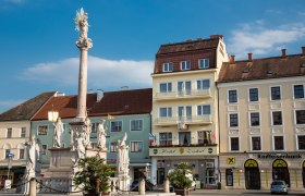 A square with a column and statues, surrounded by buildings, including the Hotel Zentral.